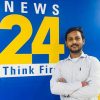 Man with folded arms posing in front of a blue wall that shows a 'News 24' sign and 'Think First' text behind him.