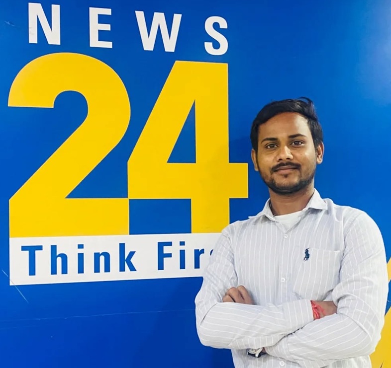 Man with folded arms posing in front of a blue wall that shows a 'News 24' sign and 'Think First' text behind him.