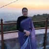 Woman in a purple sari standing on a terrace at sunset with the sea and hills in the background.