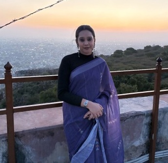 Woman in a purple sari standing on a terrace at sunset with the sea and hills in the background.
