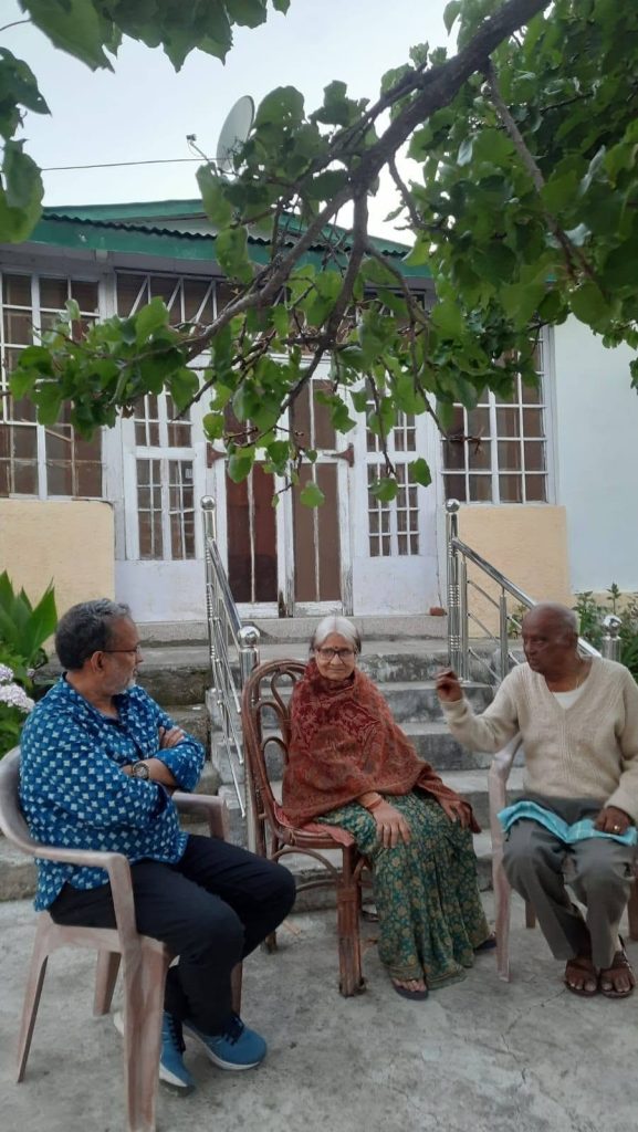 shrivastava_pahad | No. 1 Indian Media News Portal Three elderly adults chatting in a courtyard outside a house, seated on chairs under a leafy tree; left man in a blue patterned shirt, center woman in a shawl, right man in a beige cardigan.