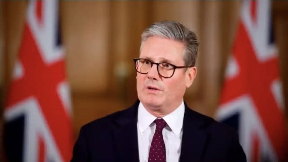 stormer | No. 1 Indian Media News Portal Man in a dark suit and glasses speaks at a press conference with Union Jack flags in the background, formal setting.