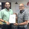 Two men in an office exchange a document, smiling at the camera during a formal handover.