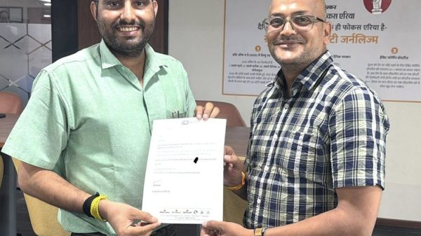 Two men in an office exchange a document, smiling at the camera during a formal handover.