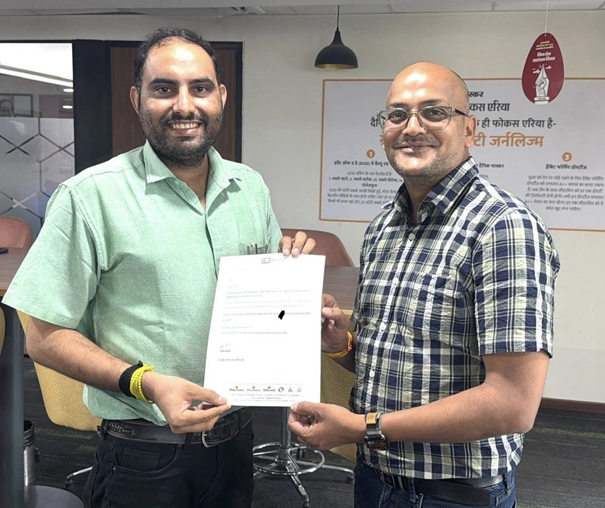 Two men in an office exchange a document, smiling at the camera during a formal handover.