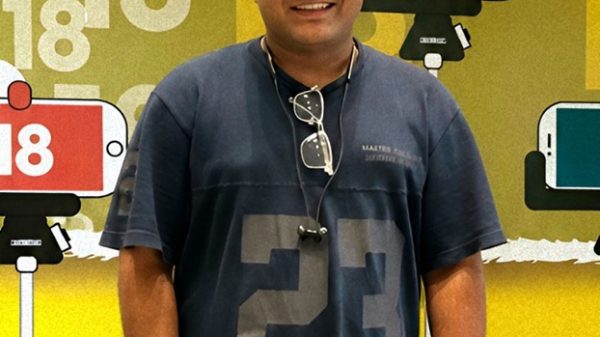 Smiling man in a dark blue T-shirt with the number 23, cap, and sunglasses hanging from his neck, standing in front of a colorful backdrop with large 18s and phone props; a small bird sits on a red phone sign.
