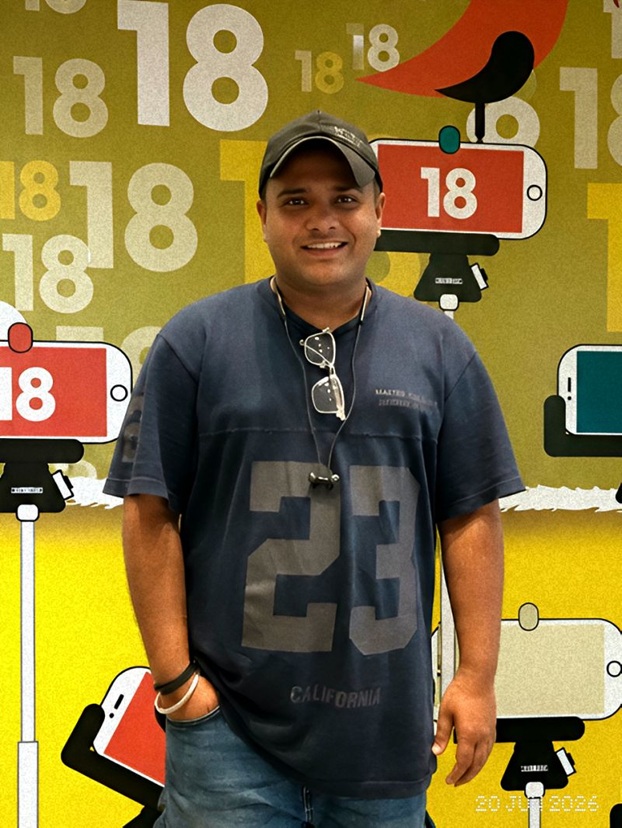 Smiling man in a dark blue T-shirt with the number 23, cap, and sunglasses hanging from his neck, standing in front of a colorful backdrop with large 18s and phone props; a small bird sits on a red phone sign.