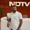 Man wearing a white polo stands at a rounded white reception desk with an orange wall and the NDTV logo in the background, posing for the camera.