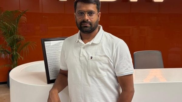 Man wearing a white polo stands at a rounded white reception desk with an orange wall and the NDTV logo in the background, posing for the camera.