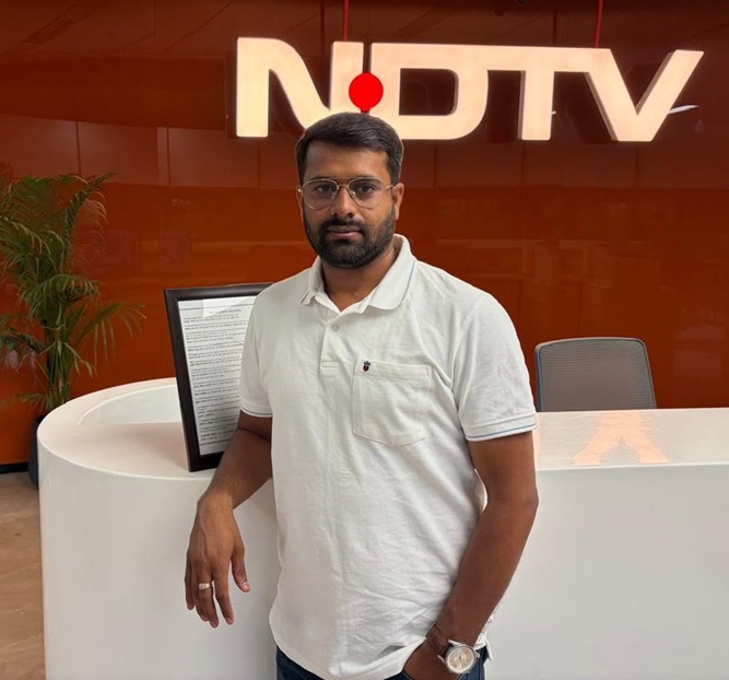 Man wearing a white polo stands at a rounded white reception desk with an orange wall and the NDTV logo in the background, posing for the camera.