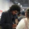 Man with curly hair and glasses in a blazer signs a notebook while holding two books at a busy indoor event.