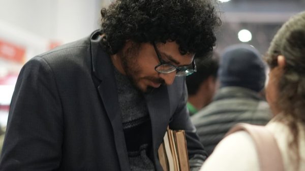 Man with curly hair and glasses in a blazer signs a notebook while holding two books at a busy indoor event.