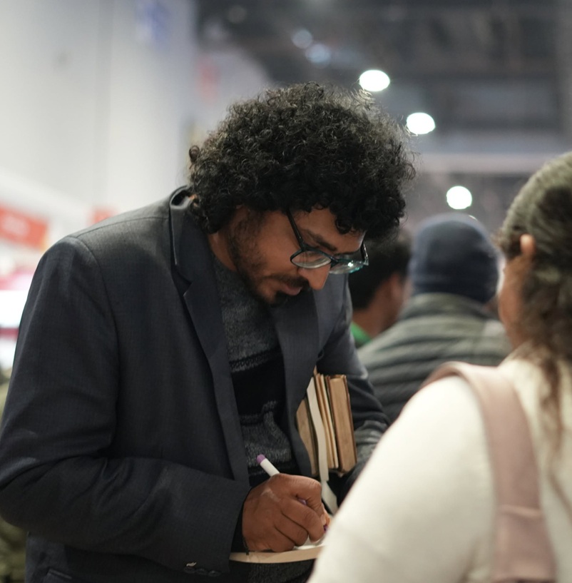 sushobhit | No. 1 Indian Media News Portal Man with curly hair and glasses in a blazer signs a notebook while holding two books at a busy indoor event.