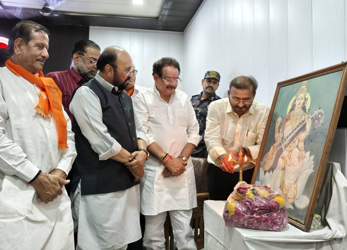 Group of men in traditional Indian attire paying respects to a framed painting of a goddess, with a lit lamp and flower garlands nearby.