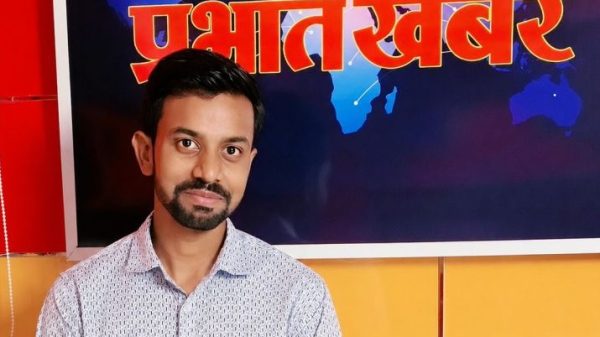 Portrait of a man with short dark hair and a beard, wearing a light patterned shirt, standing in a TV news studio backdrop.