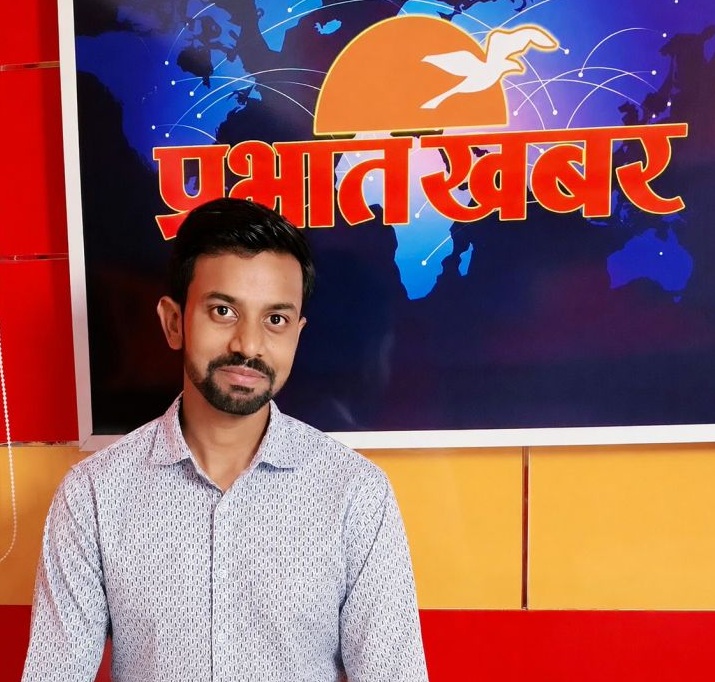 Portrait of a man with short dark hair and a beard, wearing a light patterned shirt, standing in a TV news studio backdrop.