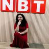 Woman in a red dress kneels on a wooden floor, resting her chin on her hand, in front of a beige wall with large red letters N B T above her