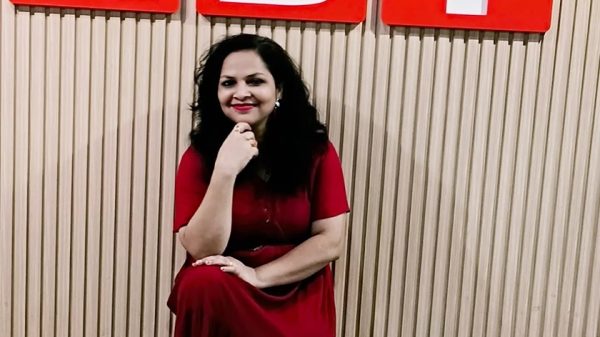 Woman in a red dress kneels on a wooden floor, resting her chin on her hand, in front of a beige wall with large red letters N B T above her