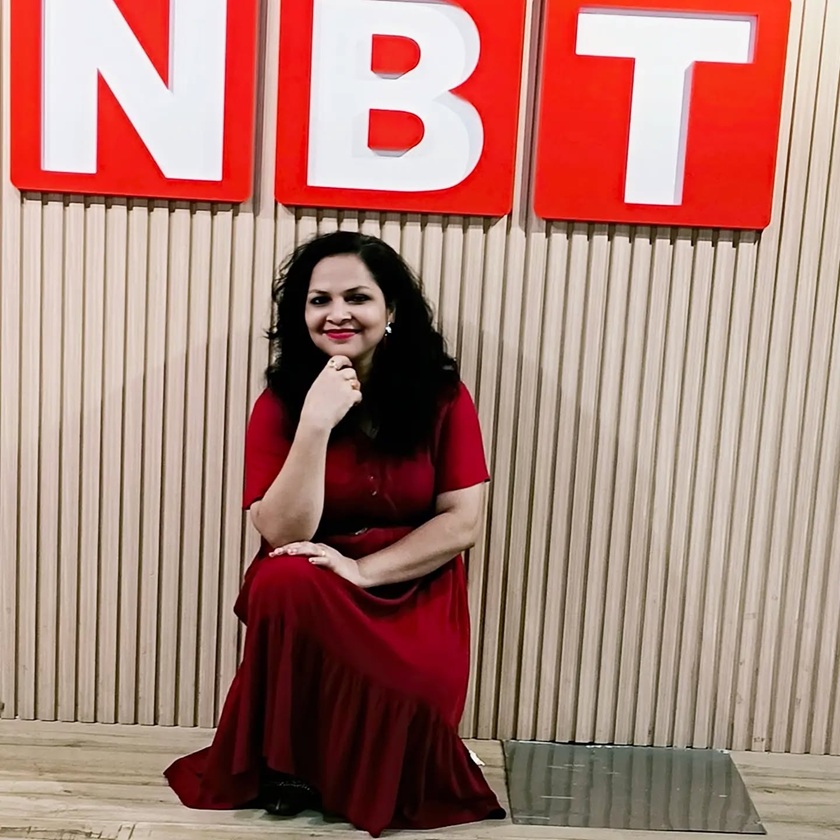 Woman in a red dress kneels on a wooden floor, resting her chin on her hand, in front of a beige wall with large red letters N B T above her