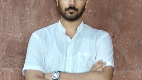Man with arms crossed, wearing a white short-sleeve shirt, standing against a brown textured wall.