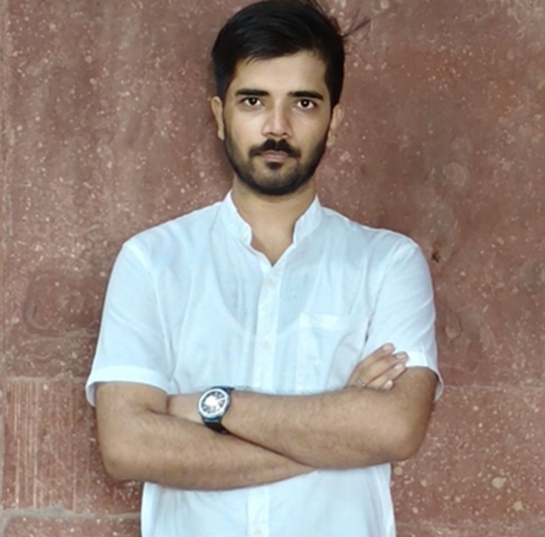 Man with arms crossed, wearing a white short-sleeve shirt, standing against a brown textured wall.