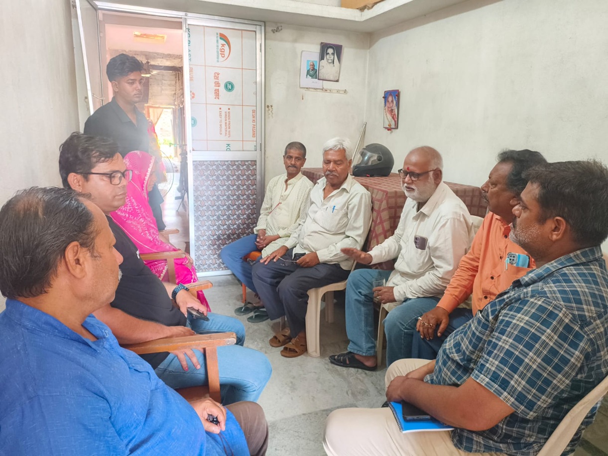 Group of men and one woman seated in a circle in a small room, having a discussion; a man stands by the doorway in the background.