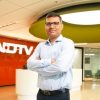 Portrait of a man in a light blue shirt standing with arms crossed in a modern NDTV newsroom lobby.