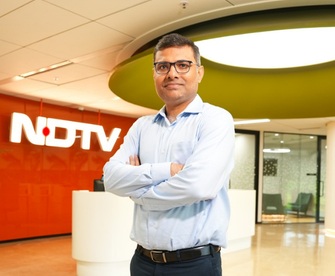 Portrait of a man in a light blue shirt standing with arms crossed in a modern NDTV newsroom lobby.