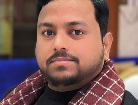 Close-up portrait of a man with short black hair and a burgundy check scarf looking at the camera.
