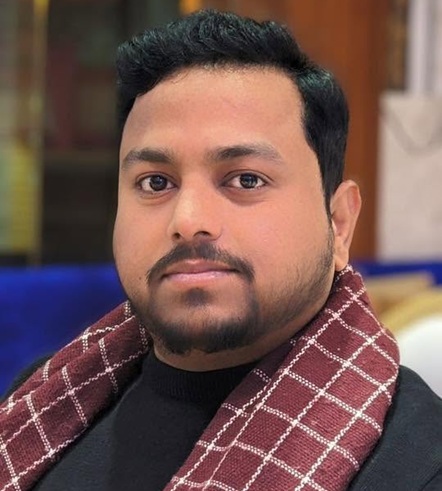 Close-up portrait of a man with short black hair and a burgundy check scarf looking at the camera.