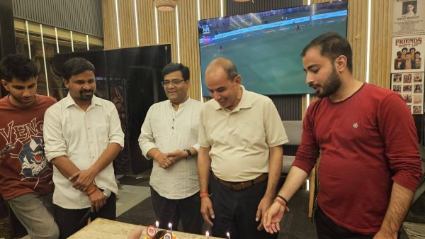 Group of five men gathered around a table with two birthday cakes and lit candles, celebrating in a modern room with a mounted TV showing a sports game in the background, warm lighting above.