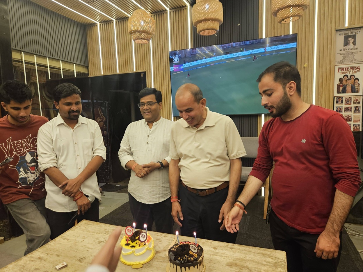 Group of five men gathered around a table with two birthday cakes and lit candles, celebrating in a modern room with a mounted TV showing a sports game in the background, warm lighting above.