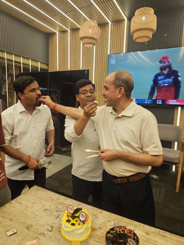 Group of men in white shirts sharing cake at a celebration indoors, with a cake on the table in front of them.