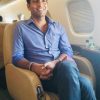 Smiling man in a blue button-down shirt sits in an airplane seat with a window and tray area nearby.