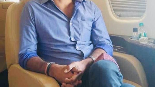 Smiling man in a blue button-down shirt sits in an airplane seat with a window and tray area nearby.