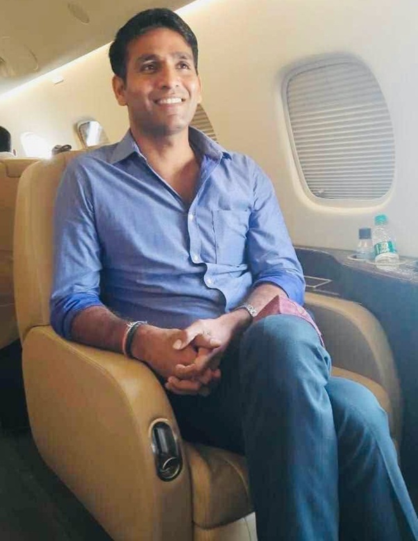 Smiling man in a blue button-down shirt sits in an airplane seat with a window and tray area nearby.