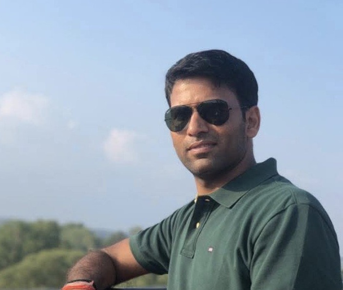 Man wearing dark sunglasses and a green polo shirt leaning on a railing outdoors on a sunny day with a blue sky behind him, suggesting a casual portrait.