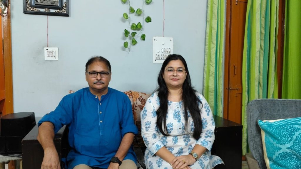 A man in a blue kurta and a woman in a blue-printed white dress sit together on a sofa in a living room with green curtains and plants behind them.
