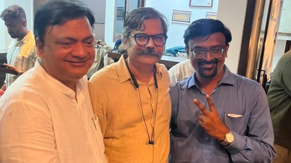 Three men pose together indoors, smiling; left in white shirt, center in yellow shirt with glasses, right in blue shirt making a peace sign.