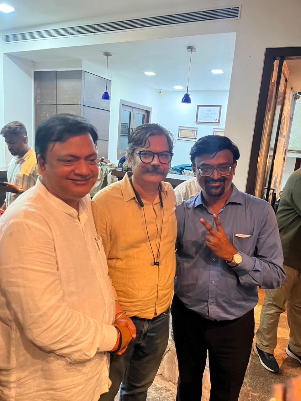 Three men pose together indoors, smiling; left in white shirt, center in yellow shirt with glasses, right in blue shirt making a peace sign.