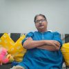 Man in a blue kurta sits on a sofa with yellow patterned cushions, arms crossed, wearing glasses; a colorful bookshelf and a plush toy are behind him.