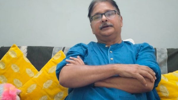 Man in a blue kurta sits on a sofa with yellow patterned cushions, arms crossed, wearing glasses; a colorful bookshelf and a plush toy are behind him.