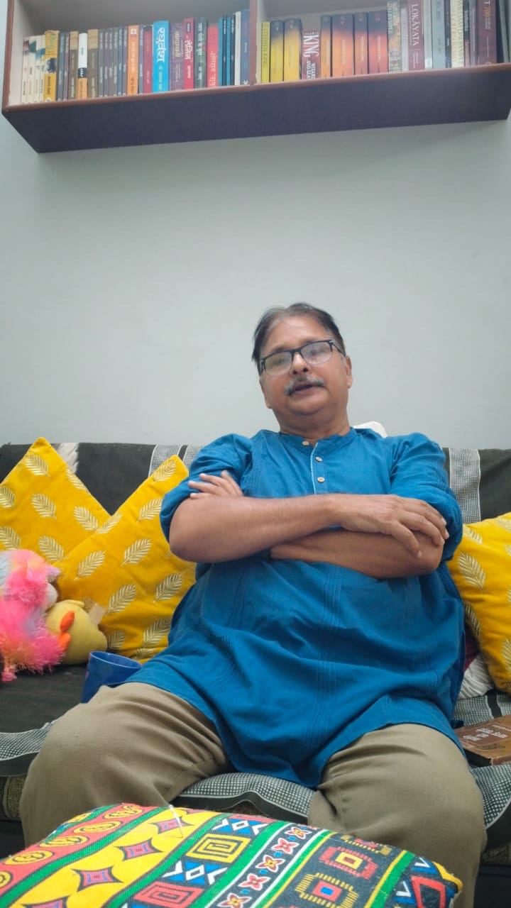 Man in a blue kurta sits on a sofa with yellow patterned cushions, arms crossed, wearing glasses; a colorful bookshelf and a plush toy are behind him.