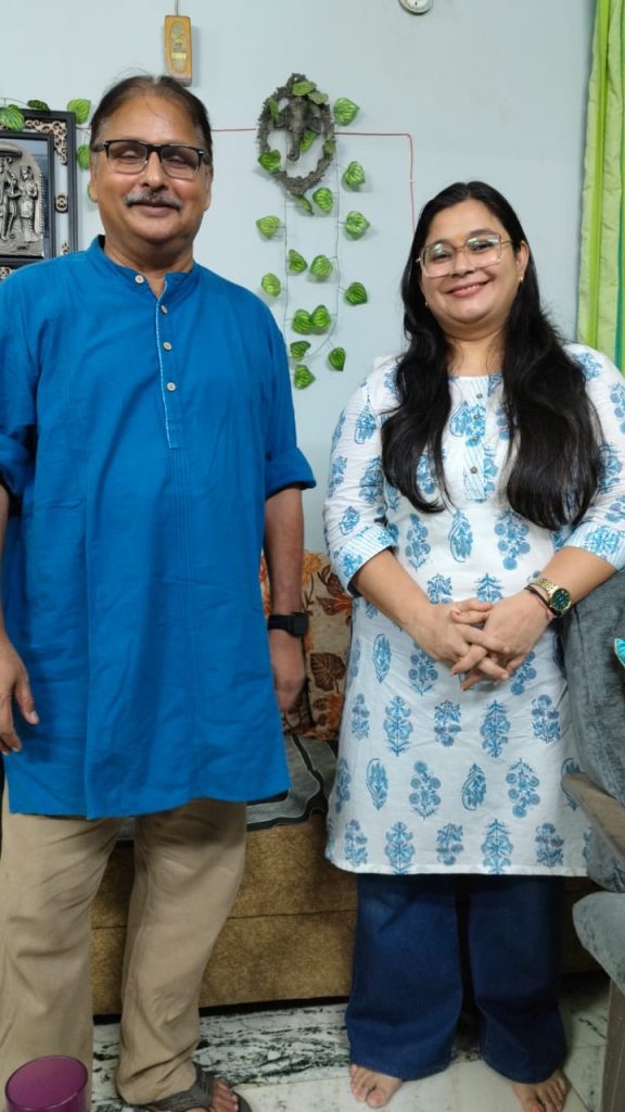 Man in a blue kurta and a woman in a white and blue patterned kurta pose together indoors, smiling at the camera.
