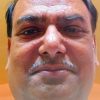 Close-up of a middle-aged man's face with a mustache, looking at the camera against an orange wall.