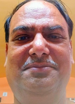 Close-up of a middle-aged man's face with a mustache, looking at the camera against an orange wall.