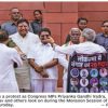 Group of Indian politicians protest outside Parliament; central figure in white shirts greets supporters while a banner with faces is held to the right.