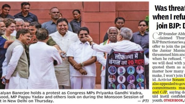 Group of Indian politicians protest outside Parliament; central figure in white shirts greets supporters while a banner with faces is held to the right.