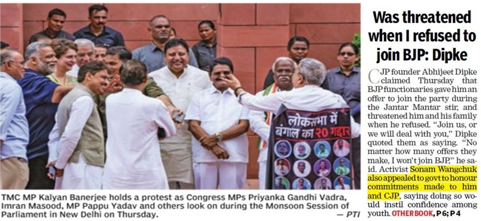 Group of Indian politicians protest outside Parliament; central figure in white shirts greets supporters while a banner with faces is held to the right.