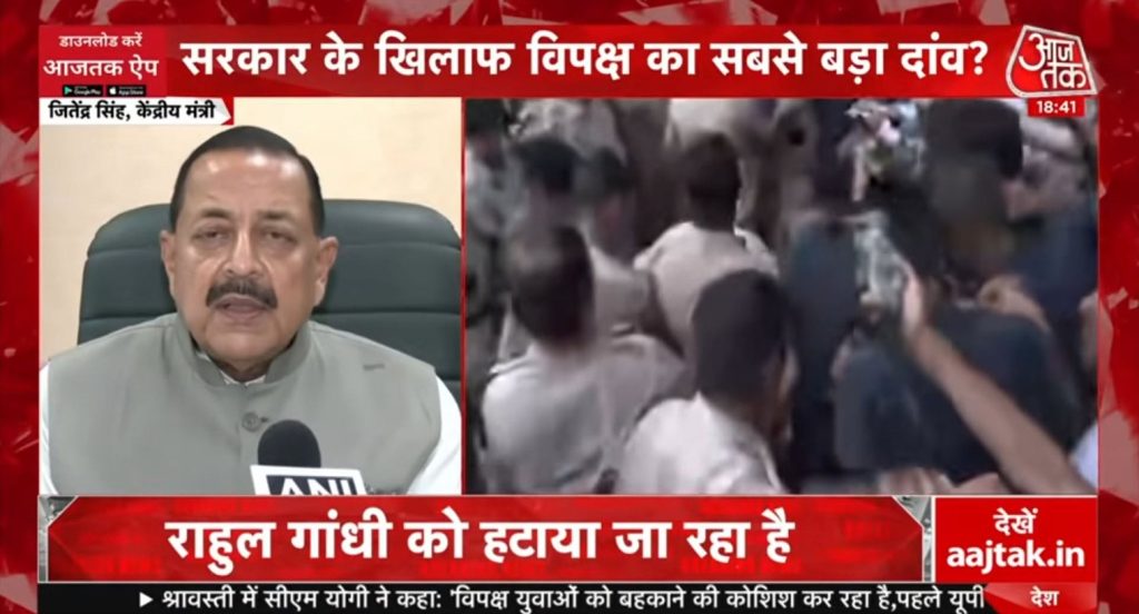 aajtak | No. 1 Indian Media News Portal Split-screen news broadcast: left panel shows a man in a beige Nehru jacket speaking into a microphone, right panel shows a blurred crowd in a busy scene; Aaj Tak branding and Hindi news banners framing the screen.
